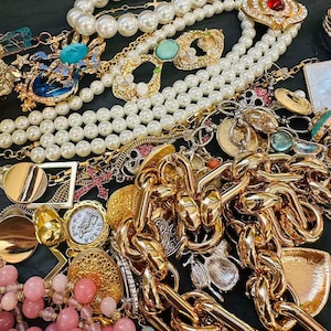 K&ouml;nnte beinhalten: Eine Sammlung von Vintage-Schmuck, darunter Goldketten, Perlenketten und verzierte Broschen. Das Sortiment umfasst verschiedene Farben, darunter Gold, Wei&szlig;, Rosa und T&uuml;rkis. Die Schmuckst&uuml;cke sind eng beieinander angeordnet und zeigen eine Vielzahl von Stilen und Designs.