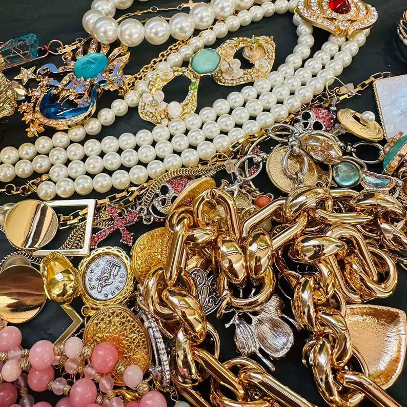 K&ouml;nnte beinhalten: Eine Sammlung von Vintage-Schmuck, darunter Goldketten, Perlenketten und verzierte Broschen. Das Sortiment umfasst verschiedene Farben, darunter Gold, Wei&szlig;, Rosa und T&uuml;rkis. Die Schmuckst&uuml;cke sind eng beieinander angeordnet und zeigen eine Vielzahl von Stilen und Designs.