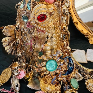 K&ouml;nnte beinhalten: Eine Sammlung von verziertem, goldfarbenem Schmuck, darunter Halsketten, Anh&auml;nger und Broschen. Die St&uuml;cke weisen Edelsteine, Perlen und Himmelsmotive wie Sonnen und Monde auf. Ein dekorativer Spiegelrahmen ist sichtbar.