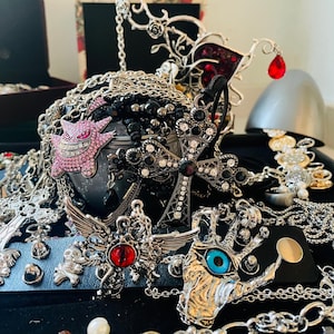 Puede incluir: Una colección de joyas de estilo gótico, que incluye collares, colgantes y pulseras. Las piezas presentan cruces, alas y otros diseños ornamentados, con acentos en negro, plata y rojo. También hay un personaje de dibujos animados rosa y enjoyado.