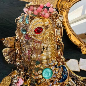 K&ouml;nnte beinhalten: Eine verzierte Ausstellung von Vintage-Schmuck, darunter Halsketten, Armb&auml;nder und Broschen. Die Kollektion umfasst goldfarbene Ketten, Perlenstr&auml;nge und Anh&auml;nger mit roten, blauen und t&uuml;rkisfarbenen Akzenten. Im Hintergrund ist ein goldgerahmter Spiegel zu sehen.