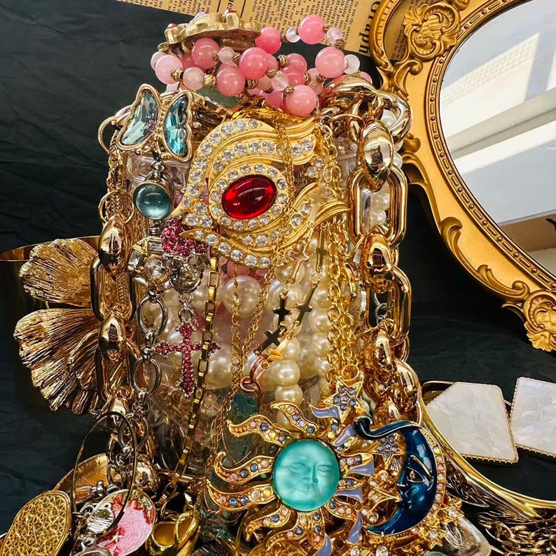 K&ouml;nnte beinhalten: Eine verzierte Ausstellung von Vintage-Schmuck, darunter Halsketten, Armb&auml;nder und Broschen. Die Kollektion umfasst goldfarbene Ketten, Perlenstr&auml;nge und Anh&auml;nger mit roten, blauen und t&uuml;rkisfarbenen Akzenten. Im Hintergrund ist ein goldgerahmter Spiegel zu sehen.