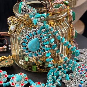 May include: A collection of turquoise and silver-toned jewelry, including necklaces, bracelets, and rings, is displayed in and around a glass jar. The jewelry features various designs, including beaded strands and ornate pendants, with red and silver accents.