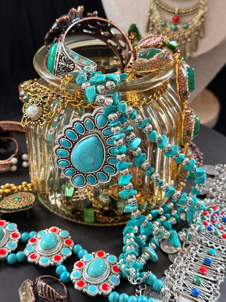 May include: A collection of turquoise and silver-toned jewelry, including necklaces, bracelets, and rings, is displayed in and around a glass jar. The jewelry features various designs, including beaded strands and ornate pendants, with red and silver accents.