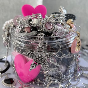 May include: A clear jar overflowing with various silver-toned jewelry, including rings, necklaces, and charms. A large, bright pink heart and a star-shaped pendant with a skull design are prominent. Other jewelry pieces include earrings and bracelets.