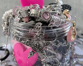 May include: A clear jar overflowing with various silver-toned jewelry, including rings, necklaces, and charms. A large, bright pink heart and a star-shaped pendant with a skull design are prominent. Other jewelry pieces include earrings and bracelets.