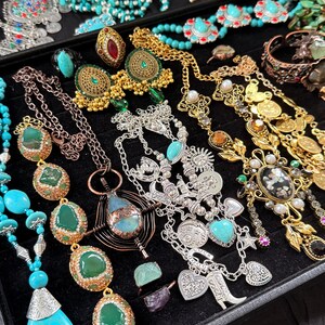 May include: A collection of jewelry displayed on a black tray. The assortment includes necklaces, earrings, rings, and bracelets in various styles. Turquoise, green, gold, and silver tones are prominent, with beaded and ornate designs. Some pieces feature gemstones and charms.