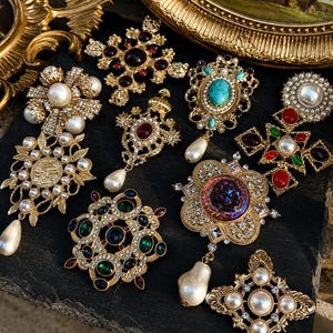 May include: A collection of gold-tone metal brooches featuring various designs with faux pearls, colorful cabochon stones, and clear crystals. The pieces include cross shapes, floral motifs, and pendants with dangling teardrop-shaped faux pearls.