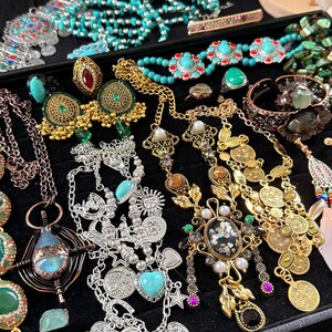 May include: A collection of various necklaces, earrings, rings, and bracelets. The jewelry features turquoise, gold, silver, and other colorful gemstones. The pieces are displayed on a black surface, showcasing a bohemian or vintage style.
