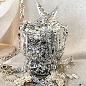 May include: A clear, ribbed glass container filled with an assortment of silver-toned jewelry, including chains, rhinestones, and white pearls. A large, textured silver starfish ornament rests on top, while gold-toned butterfly charms surround the base.