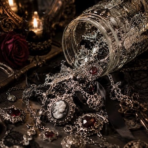 May include: A clear glass jar tipped on its side, spilling out an assortment of silver-toned jewellery. The collection includes necklaces with dark red faceted stones, a cameo pendant featuring a white profile, and intricate metal filigree charms.