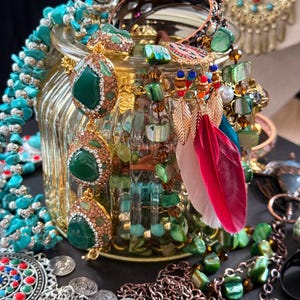 May include: A collection of jewelry, including necklaces, earrings, and rings, in a variety of styles and colors. The pieces feature turquoise, green, and gold accents, with some adorned with gemstones and feathers. The jewelry is displayed in and around a decorative glass jar.
