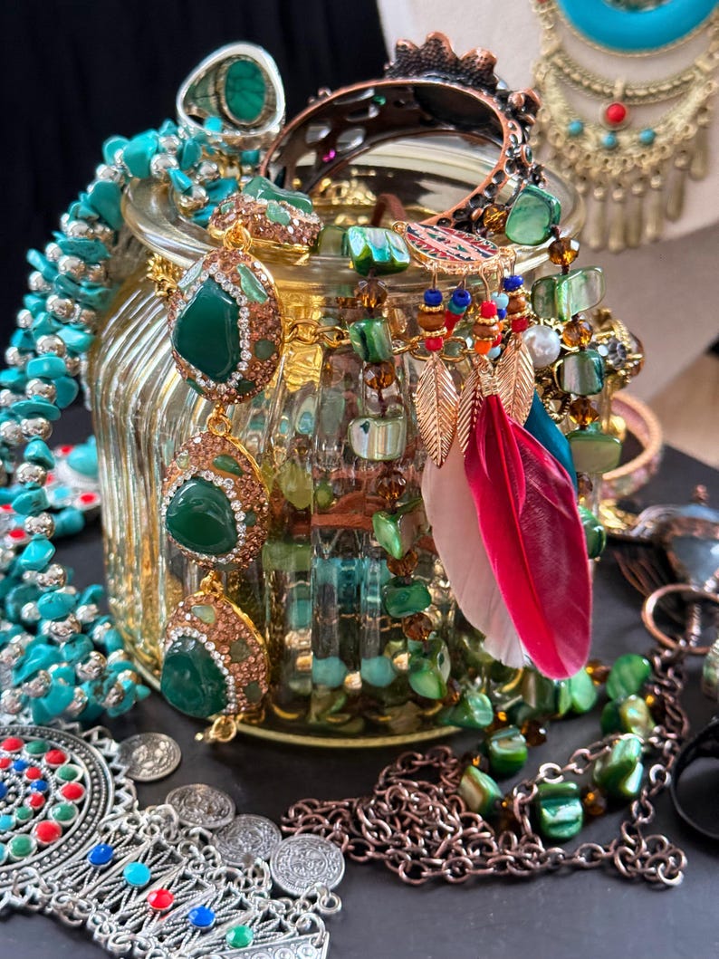 May include: A collection of jewelry, including necklaces, earrings, and rings, in a variety of styles and colors. The pieces feature turquoise, green, and gold accents, with some adorned with gemstones and feathers. The jewelry is displayed in and around a decorative glass jar.