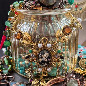 May include: A collection of ornate jewelry, including rings, necklaces, and bracelets, displayed around a glass jar. The jewelry features gold-toned metal, gemstones in various colors, and intricate designs. The jar is filled with turquoise beads.