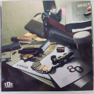 May include: A CD case featuring the album "Section.80" by Kendrick Lamar. The cover art displays a still life with a gold chain, a pipe, and a pill bottle. The album title and artist name are visible.