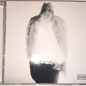 Könnte beinhalten: Eine CD-Hülle mit einem Schwarz-Weiß-Bild einer Figur mit verschwommenem, strukturiertem Aussehen. Die Figur ist vor einem weißen Hintergrund zentriert. Die Worte "Parental Advisory Explicit Content" sind in Schwarz gedruckt.