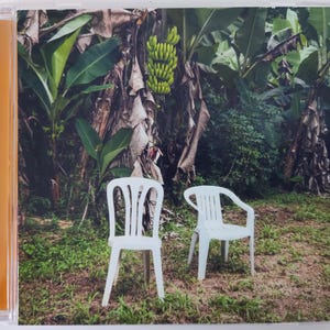 Puede incluir: Una caja de CD con una fotografía de dos sillas de plástico blancas en un entorno tropical. El fondo incluye exuberante follaje verde, plataneros y un racimo de plátanos verdes. La caja tiene un lomo naranja.