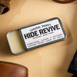 May include: An overhead shot features leather care products and accessories. A tin of "HIDE REVIVE" leather conditioner sits open, next to a black leather glove, a brown leather boot, and a leather case. The text on the tin reads "CONDITIONS, CLEANS, & RESTORES LEATHER GOODS LARGE OR SMALL."