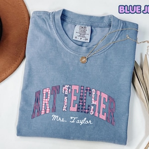 Puede incluir: Una camiseta color azul jean con cuello redondo y etiqueta de la marca Comfort Colors. La prenda presenta el texto ART TEACHER en una fuente rosa y azul con múltiples patrones, con el nombre Mrs. Taylor escrito debajo en cursiva blanca.