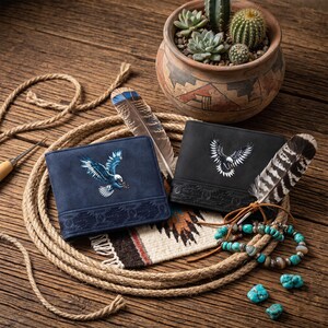 May include: Two leather wallets, one navy blue and one black, each featuring an embroidered eagle design. The wallets are displayed with feathers, a turquoise bracelet, and a small pot of succulents on a wooden surface.