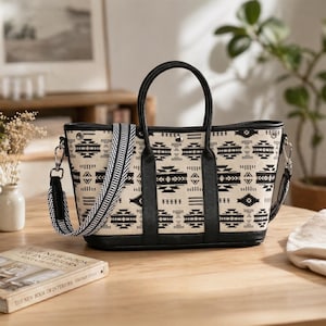 May include: A beige tote bag with a black geometric pattern and black leather handles. The bag has a detachable black and white patterned strap. The bag is sitting on a wooden surface.