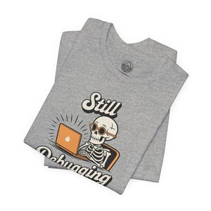 May include: A grey t-shirt featuring a skeleton wearing sunglasses and working on an orange laptop. The text "Still Debugging" is above and below the skeleton. The t-shirt is folded.