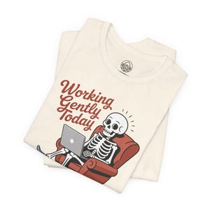 Könnte beinhalten: Cremefarbenes T-Shirt mit einem Cartoon-Skelett, das sich in einem roten Sessel entspannt und einen Laptop benutzt. Der Satz "Working Gently Today" ist in roter Schrift über dem Skelett aufgedruckt. Das T-Shirt ist gefaltet und zeigt den Kragen und ein kleines Logo.