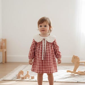 May include: A young child wearing a red and white checkered dress with a white collar and long sleeves. The dress has a tie at the neckline. The child is standing in a room with wooden toys and a rocking horse.