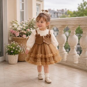 May include: A toddler dress featuring a cream-colored long-sleeve top with ruffled shoulders, paired with a brown bodice overlay that has two small bows and two white buttons. The skirt is made of layered brown tulle with ruffled trim.