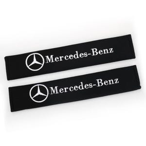 May include: Two rectangular black seat belt covers featuring the white Mercedes-Benz logo and the text Mercedes-Benz printed on the front. The covers are made of a soft fabric material and are designed to wrap around standard seat belts.
