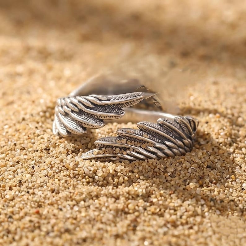 K&ouml;nnte beinhalten: Ein silberfarbener Ring mit detailliertem Fl&uuml;geldesign. Der Ring ist offen, wobei die Fl&uuml;gelspitzen voneinander weg zeigen. Der Ring liegt auf Sand.