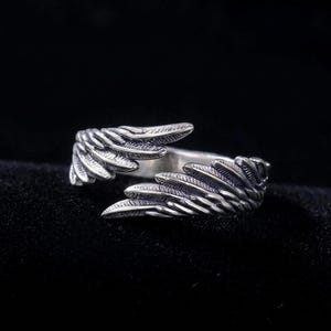 K&ouml;nnte beinhalten: Ein silberner Ring mit einem Fl&uuml;geldesign. Der Ring hat zwei Fl&uuml;gel, die sich um den Finger legen und ein offenes Design bilden. Die Fl&uuml;gel haben strukturierte Federdetails. Der Ring ist vor einem dunklen Hintergrund platziert.