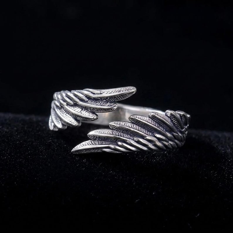 K&ouml;nnte beinhalten: Ein silberner Ring mit einem Fl&uuml;geldesign. Der Ring hat zwei Fl&uuml;gel, die sich um den Finger legen und ein offenes Design bilden. Die Fl&uuml;gel haben strukturierte Federdetails. Der Ring ist vor einem dunklen Hintergrund platziert.