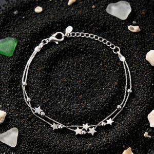May include: A silver bracelet with two strands, adorned with star and bead details. It has a lobster clasp and an adjustable chain. The jewellery is presented on a black surface, with shells and sea glass scattered around.