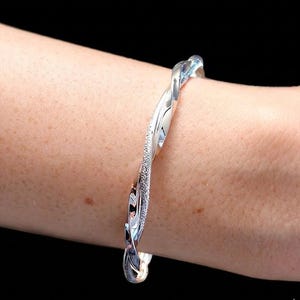 Könnte beinhalten: Ein silberfarbenes, verdrehtes Armreif-Armband. Das Armband hat auf einer Seite eine strukturierte, glitzernde Oberfläche und auf der anderen Seite eine glatte, polierte Oberfläche. Das Armband wird an einem Arm getragen.