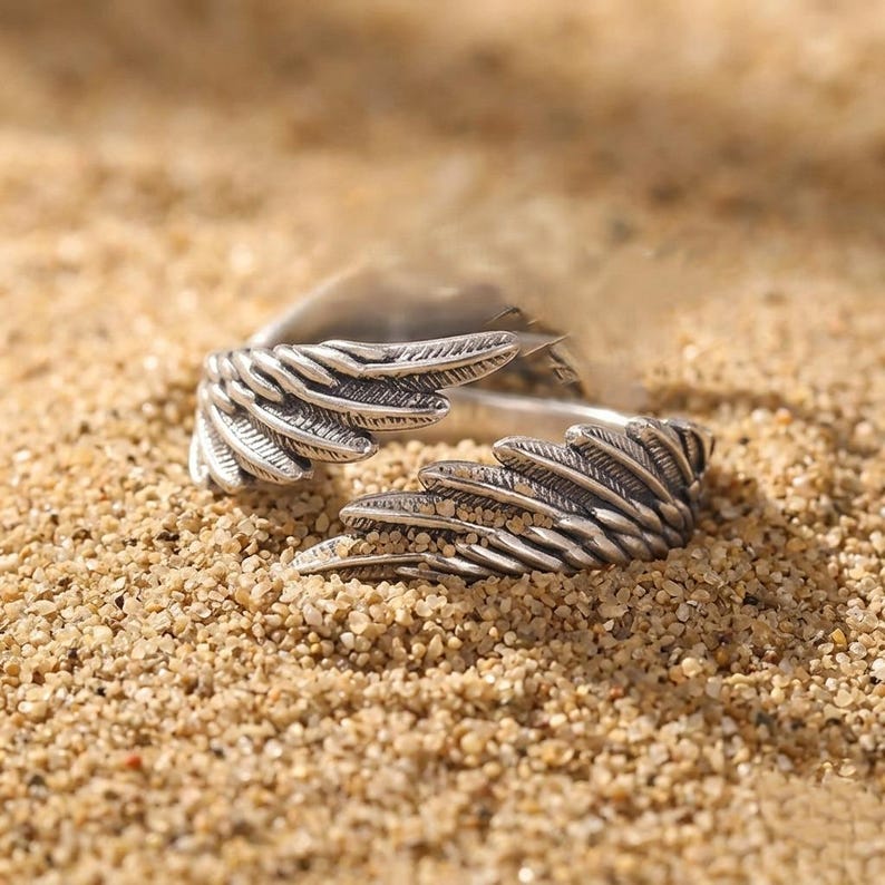K&ouml;nnte beinhalten: Ein silberfarbener Ring mit einem Fl&uuml;geldesign. Der Ring weist detaillierte Federstrukturen auf, wobei sich die Fl&uuml;gel nach au&szlig;en erstrecken. Der Ring ist vor einem sandigen Hintergrund platziert, was einen Kontrast zwischen dem Metall und den nat&uuml;rlichen Farbt&ouml;nen erzeugt.
