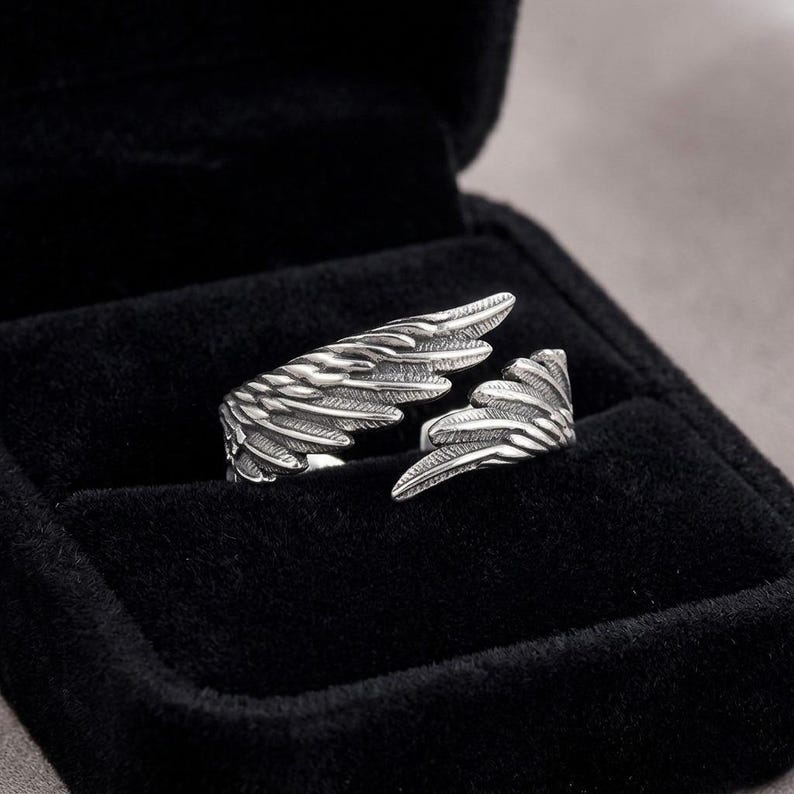 K&ouml;nnte beinhalten: Ein silberfarbener Ring in Form von Engelsfl&uuml;geln, offen in einer schwarzen Schmuckschatulle pr&auml;sentiert. Das Design des Rings weist detaillierte Federtexturen auf und schafft so ein realistisches und elegantes Accessoire. Der Ring ist ein einzigartiges Schmuckst&uuml;ck.