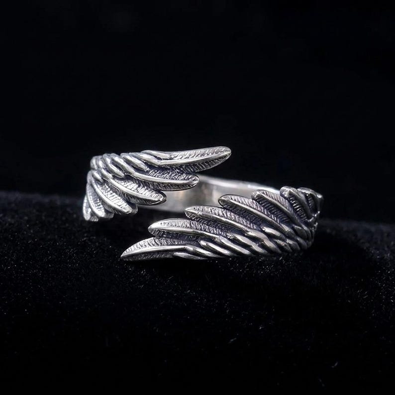 K&ouml;nnte beinhalten: Ein silberner Ring mit einem Fl&uuml;geldesign. Der Ring hat zwei stilisierte Fl&uuml;gel, die sich um den Finger legen und ein offenes Design bilden. Die Fl&uuml;gel haben strukturierte Federdetails, vor einem dunklen Hintergrund.