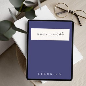 May include: A digital tablet with a dark blue cover and the words "CHOOSE A LIFE YOU Love" in a white rectangle. The word "LEARNING" is at the bottom. Gold-rimmed glasses rest on a white book, with green leaves in the background.