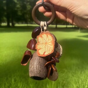 May include: A brown wooden ring keyring with a collection of natural elements. The keyring features a dried orange slice, small brown shoe-shaped charms, and other organic shapes. The background is a blurred green field.