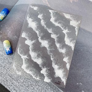 May include: Transparent nail art sticker with a white wave design on a grey backdrop. Two painted fingernails are visible, one blue with a seashell design and the other yellow and blue with a star design. The sticker sheet has the text "Pm YT5045".