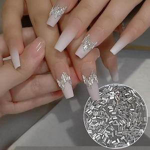 May include: Long, square-shaped nails with a white to nude ombre design, adorned with silver rectangular nail art. A small circular inset displays a pile of the silver nail art pieces. The nails are on a hand.
