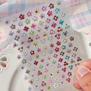 Op de afbeelding: Een transparant zelfklevend vel met talloze kleine, veelkleurige bloemvormige stickers. Elke sticker heeft een centrale kraal omringd door vijf bloemblaadjes in diverse iriserende tinten, waaronder roze, blauw, groen en paars, in een raster.
