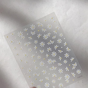 Könnte beinhalten: Transparente Nagelkunst-Aufkleberbogen mit einem Muster aus weißen Gänseblümchen mit gelben Zentren. Der Bogen wird von Fingern gehalten. Der Text "YT4978" ist in der Ecke sichtbar. Der Hintergrund ist hellgrau.