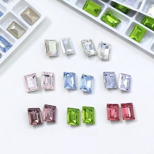 May include: Assortment of rectangular, faceted gemstones in various colors, including pink, blue, green, and clear. The gems are arranged on a white surface, some in trays. Ideal for jewelry making or craft projects.