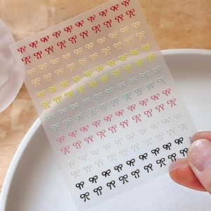 Puede incluir: Lámina transparente de pegatinas para uñas con filas de pequeños lazos en varios colores, incluyendo rojo, amarillo, rosa, blanco y negro. Los lazos están espaciados uniformemente, creando un patrón delicado para nail art.