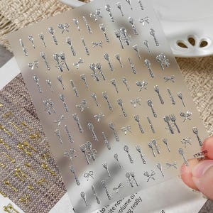 May include: A clear adhesive sheet featuring numerous small, metallic silver nail art stickers shaped like forks, spoons, knives, and bows. The bottom right corner of the sheet displays the printed text YT8299 SiPro in a small black font.