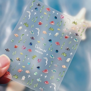 May include: A clear sheet of adhesive nail decals featuring various colourful cartoon fish, crabs, squids, eels, seaweed, and coral motifs. The sheet includes small white cross symbols and the printed text Cartoon fish YTB135Te.