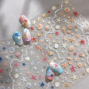 May include: Transparent sheet of nail art stickers with marine designs. Includes starfish, seashells, and other ocean elements in white, red, and gold. Several manicured nails are decorated with similar designs.