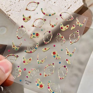May include: A sheet of adhesive nail art stickers featuring gold-toned melting clock designs, Roman numerals, and small embedded multicolored rhinestones in red, blue, and green. The sheet includes the printed text YT7594 Pro in the bottom corner.
